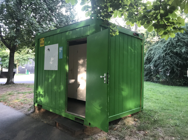 Eine grünne portable Toilette mit einer Tür und einer angebrachten Platte steht in einem Park umgeben von Bäumen und einer Bank, mit dem Himmel im Hintergrund.