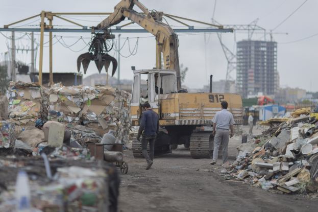 Eine Gruppe von Männern, die eine Schotterstraße entlanggehen, neben einem Haufen Müll mit einem Bagger im Hintergrund, Gebäude, Bäume und Strommasten in der Umgebung.
