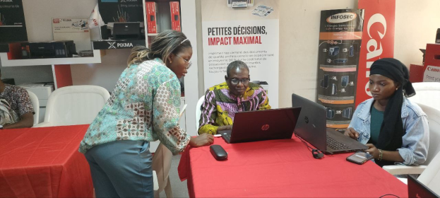Eine Gruppe von Menschen sitzt an einem Tisch mit Laptops und Mobiltelefonen auf einer internationalen Geschäftsmesse in Senegal, mit Bannern, Schränken und einer Uhr im Hintergrund.