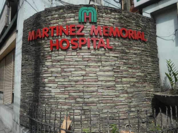 Martinez Memorial Hospital in Manila, Philippines, a multi-story building with windows, shutters, and a name board, surrounded by a metal fence and potted plants, under a cloudy sky.