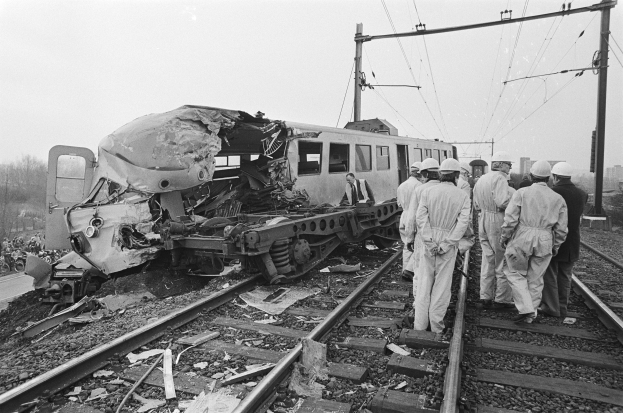 Ein Schwarz-Weiß-Bild einer Zugkollision mit Menschen in Helmen, die drumherum stehen, Strommasten, Bäumen, Gebäuden und einem klaren Himmel im Hintergrund.