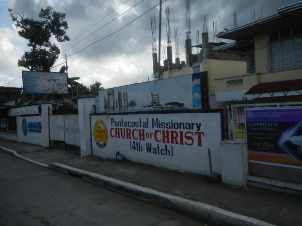 Die Pfingstmissionarische Kirche Christi 14. Stunde steht mit einer Straße im Vordergrund, einer Wand mit Bannern und Text, Gebäuden, Bäumen, Pfählen, Drähten und einem bewölkten Himmel im Hintergrund.