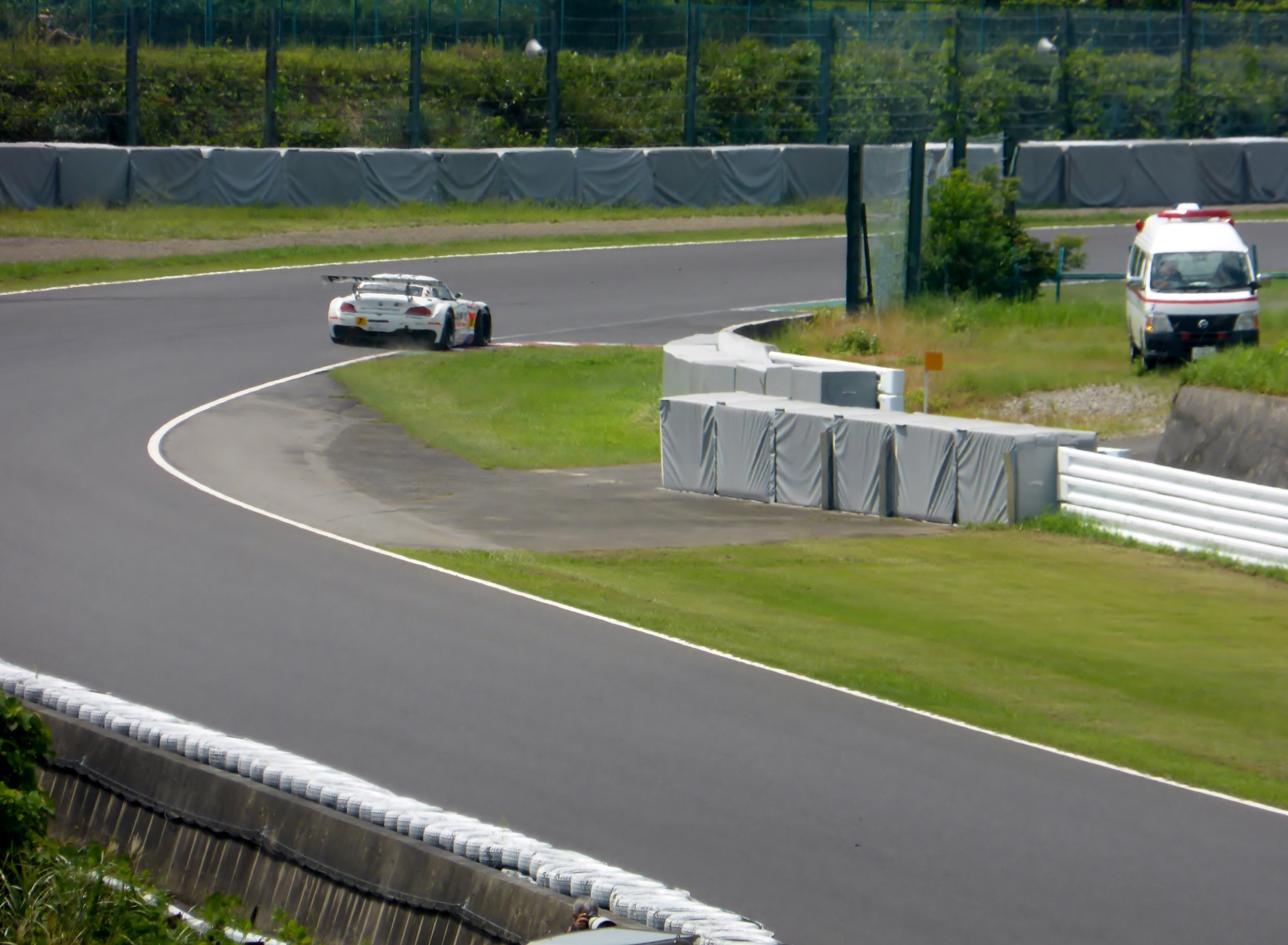 Ein Rennwagen fährt auf einer Rennstrecke, im Hintergrund ist ein Polizeiauto zu sehen, umgeben von Gras, Pflanzen, Bäumen und einem Zaun, und auf der rechten Seite des Bildes befindet sich ein Fahrzeug.