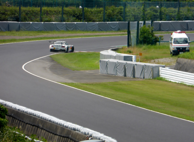 Ein Rennwagen fährt auf einer Rennstrecke, im Hintergrund ist ein Polizeiauto zu sehen, umgeben von Gras, Pflanzen, Bäumen und einem Zaun, und auf der rechten Seite des Bildes befindet sich ein Fahrzeug.