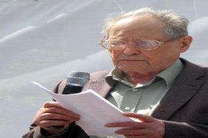 Ein älterer Mann im Anzug und mit Brille hält ein Mikrofon und Papier und sieht ernst aus.