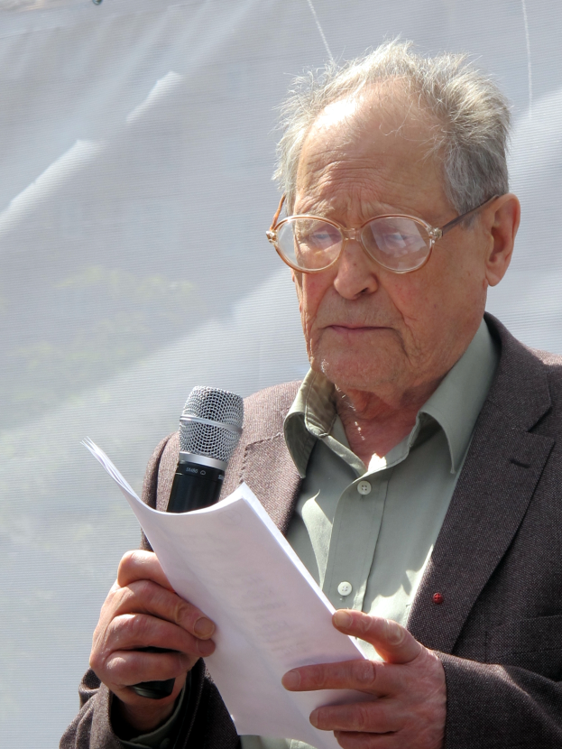 Ein älterer Mann im Anzug und mit Brille hält ein Mikrofon und Papier und sieht ernst aus.