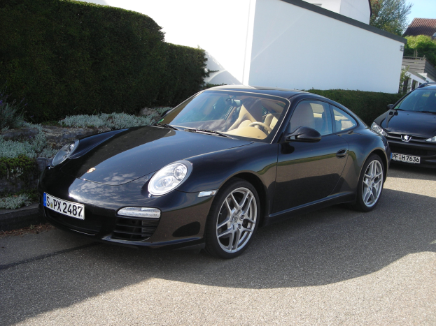 Zwei schwarze Porsche 911 vor einem weißen Haus geparkt, umgeben von üppiger grüner Vegetation und Bäumen, mit einem hellblauen Himmel im Hintergrund.