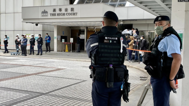 Zwei Polizisten in Uniform, Mützen und Masken stehen vor einem Gebäude am Hong Kong International Airport, umgeben von Menschen, einige halten Kameras, mit Stangen und Bändern im Hintergrund.