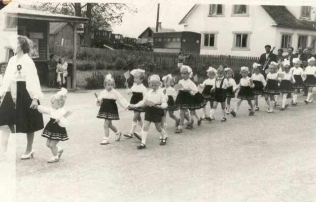 Eine Gruppe von Kindern in Schuluniformen, die eine Straße entlanggehen, mit einer Frau auf der linken Seite und im Hintergrund Gebäude, Bäume, Pflanzen, ein Zaun, Pfosten und einen klaren Himmel, alles in Schwarz-Weiß dargestellt.