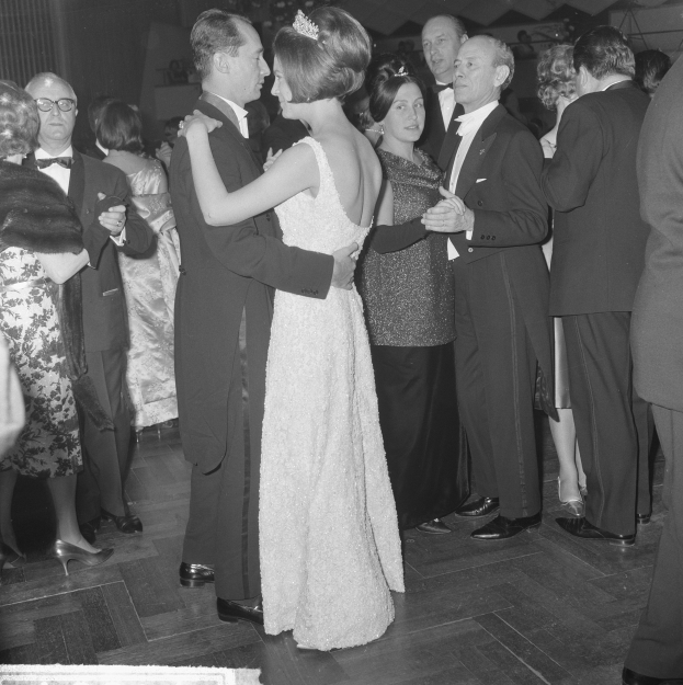Ein Schwarz-Weiß-Foto eines Paares in Abendgarderobe auf einer Tanzfläche, umgeben von einer Gruppe von Menschen, die stehen.