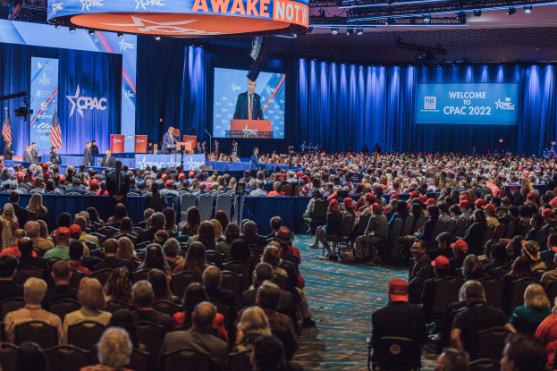 Eine große Menschenmenge sitzt auf Stühlen, die einer Bühne zugewandt sind, mit ein paar Menschen, die darauf stehen, blauen Vorhängen und einem Bildschirm im Hintergrund und einer orangefarbenen Tafel oben, die das CPAC 2020-Ereignis am 25. Oktober anzeigt.