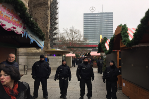 Eine Gruppe von Polizisten in schwarzen Uniformen und Mützen steht vor einem geschmützten Weihnachtsmarkt in Berlin, mit festlichen Dekoelementen, Gebäuden, Bäumen und einem klaren blauen Himmel im Hintergrund; ein Schild mit Text ist links sichtbar.