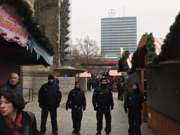 Eine Gruppe von Polizisten in schwarzen Uniformen und Mützen steht vor einem geschmützten Weihnachtsmarkt in Berlin, mit festlichen Dekoelementen, Gebäuden, Bäumen und einem klaren blauen Himmel im Hintergrund; ein Schild mit Text ist links sichtbar.