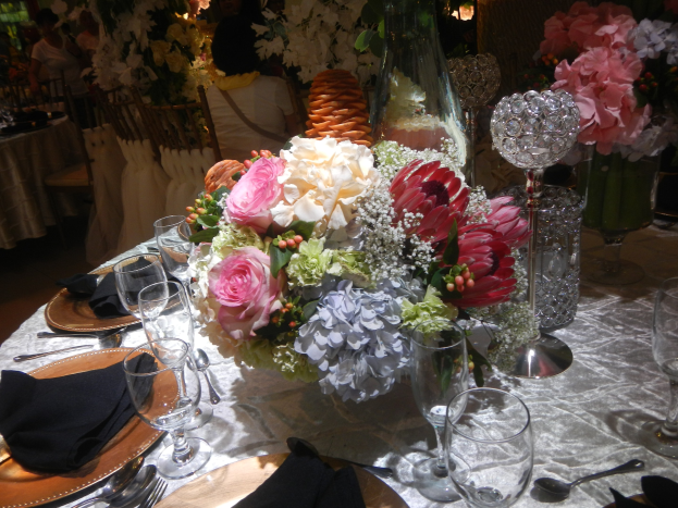 Ein formell gedeckter Esstisch mit Tellern, Besteck, Gläsern, Servietten und Blumenvasen, mit einem floralen Zentrum und im Hintergrund sitzende Gäste.