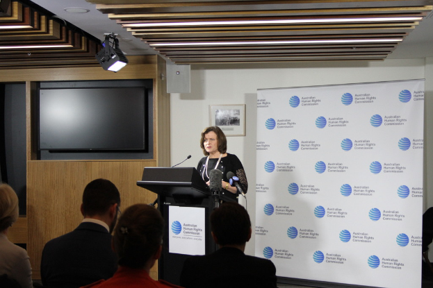 Eine Frau steht auf einer Bühne und spricht in ein Mikrofon, vor ihr sitzen Zuschauer, an der Wand hinter ihr eine Fahne, ein Fernseher an der Holzwand und Lampen an der Decke.