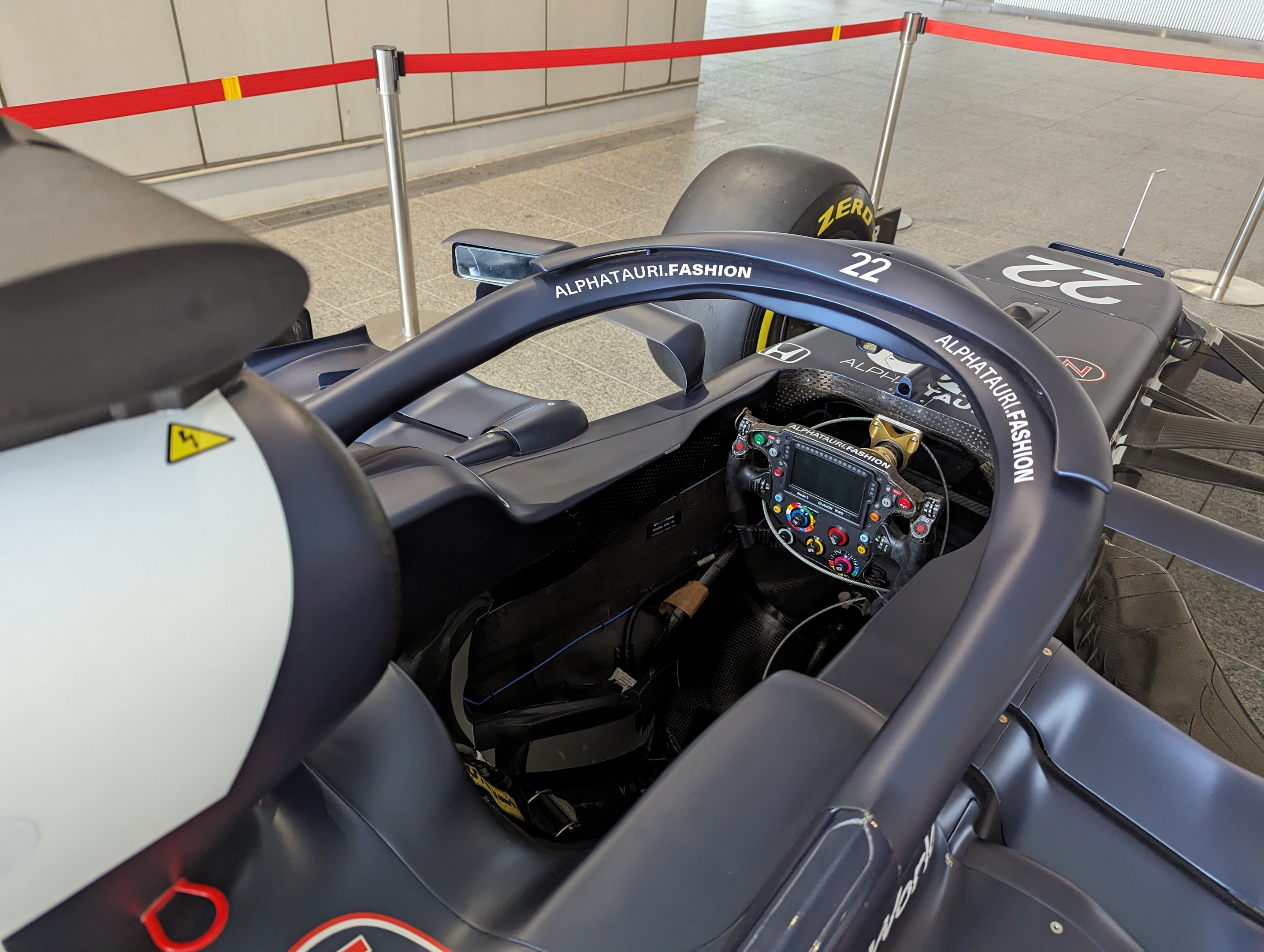 Cockpit eines Formel-E-Autos in einer Ausstellung, Lenkrad im Vordergrund, Wand und gesäumte Stangen im Hintergrund.