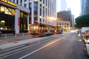 Eine Seilbahn fährt entlang einer Straße in San Francisco, mit Fahrzeugen, einem Fußgänger und einem geparkten Van im Vordergrund; Gebäude, Bäume und Himmel bilden den Hintergrund.