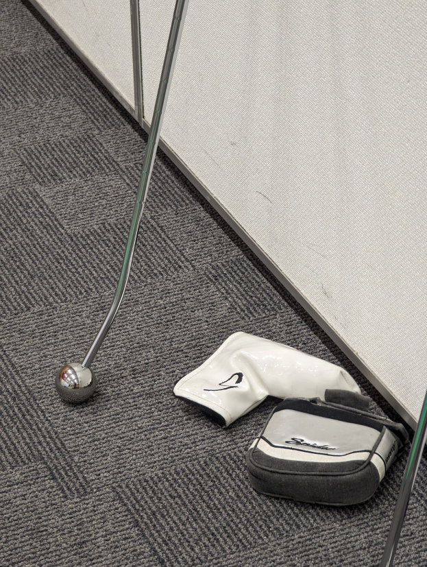 Ein silberner Golfschläger liegt auf einem Teppichboden neben einer schwarzen Golftasche mit einem weißen Logo und einem Golfschläger, an einer hellgrauen Wand mit sichtbaren Kratzern und Dellen.