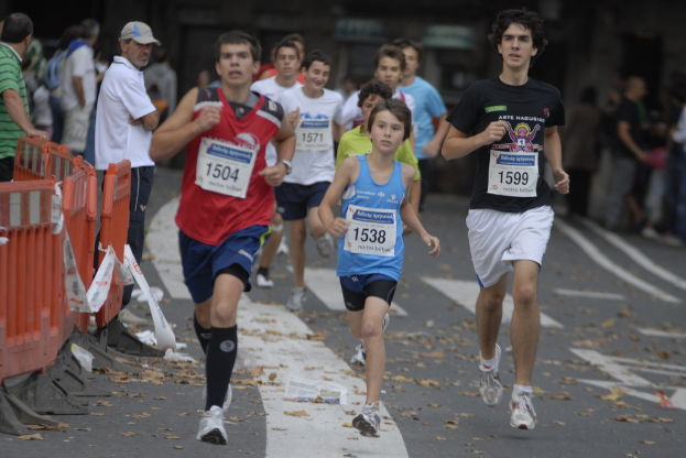 Läufer auf einer Straße mit Zuschauern auf der linken Seite hinter einem orangenen Geländer, einige tragen nummerierte T-Shirts.