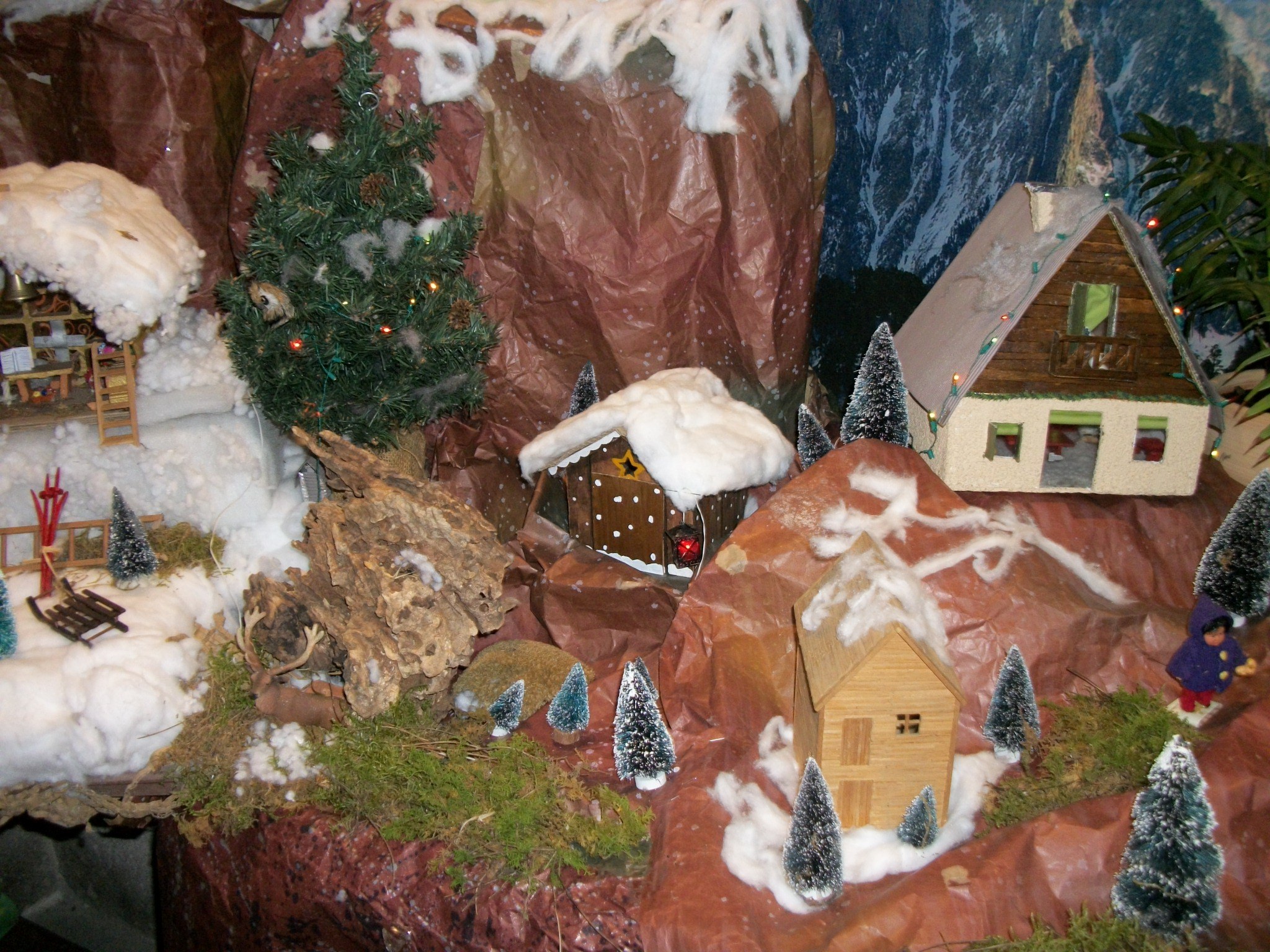 Eine Miniatur-Szene eines verschneiten Dorfes mit einem zentralen Weihnachtsbaum, umgeben von schneebedeckten Häusern, Bäumen, Pflanzen und Felsen, mit einer Hintergrundwand.