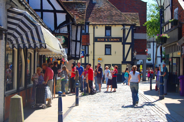 Eine Straße mit Menschen auf der linken Seite neben einem Handwagen, ein Mann und eine Frau auf der rechten Seite neben einem lila Müllcontainer, mit Gebäuden, Bäumen und Blumenkübeln im Hintergrund.