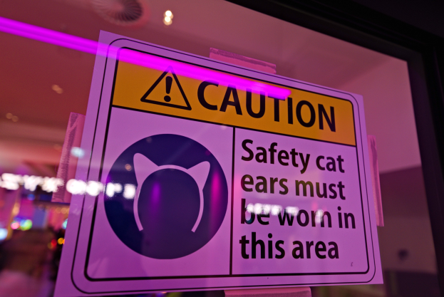 Hellgelbes Warnschild in einem Ladenschaufenster mit der Aufschrift "Vorsicht: Sicherheitskatzenohren müssen in diesem Bereich getragen werden." vor einem unscharfen Hintergrund.