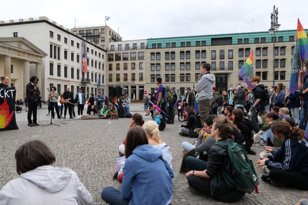 Eine Gruppe von Menschen, die auf dem Boden vor einer Menge sitzen, die Fahnen und Transparente hält, bei einer Demonstration in Berlin. Im Hintergrund sind eine Statue, ein Mikrofonständer und Gebäude zu sehen.