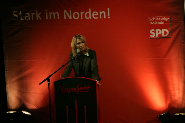 Eine Person steht neben einem Rednerpult mit einem Mikrofon auf einem Ständer, mit fokussierten Scheinwerfern und einem Banner im Hintergrund.