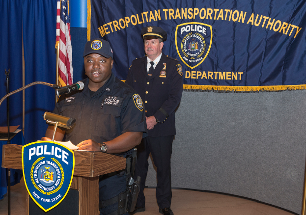 Zwei Polizeibeamte stehen vor einem Rednerpult, einer hölt Papiere und spricht in ein Mikrofon, mit einem Banner, einer Flagge und einem Tisch in der Nähe, und einer Tafel mit Symbolen und Text im Hintergrund.