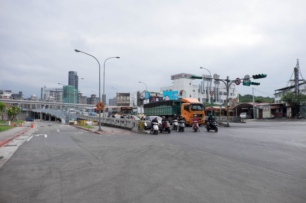 Stadtstraße mit starkem Verkehr, einschließlich eines Lastwagens und eines Autos, Laternenpfähle, Verkehrsampeln, Schilder, eine Brücke mit Geländern, Bäume, Gebäude mit Fenstern und ein bewölkter Himmel.