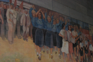 Eine Gruppe von Menschen mit blau gekleideten Mädchen in der Mitte, Musikern rechts, Kindern spielend in der rechten Ecke und Individuals mit roten Fahnen links.