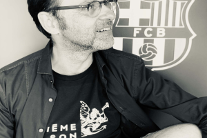 Ein Mann mit Brille und schwarzem Hemd lächelt vor einer Wand mit dem FC Barcelona-Logo, aufgenommen in Schwarz-Weiß.