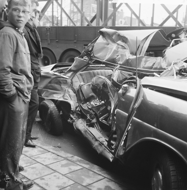 Eine schwarz-weiße Aufnahme eines auf der Seite liegenden Wagens mit mehreren Personen drumherum, einem Lastwagen und einer Brücke im Hintergrund.