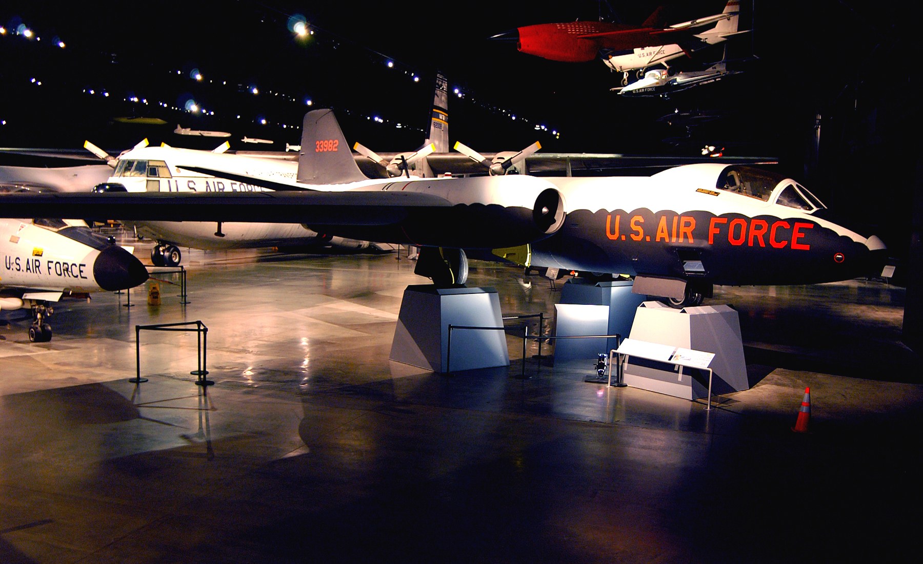 Eine Gruppe von Kampfflugzeugen in einem dunklen Museum ausgestellt, umgeben von St├Ąnden, Verkehrskegeln und anderen Gegenst├Ąnden auf dem Boden.