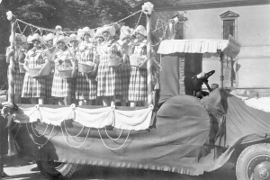 Gruppe von Frauen auf einem Truck mit Körben stehend, mit einem Gebäude und Bäumen im Hintergrund, in Schwarz-Weiß.