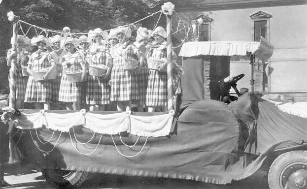 Gruppe von Frauen auf einem Truck mit Körben stehend, mit einem Gebäude und Bäumen im Hintergrund, in Schwarz-Weiß.