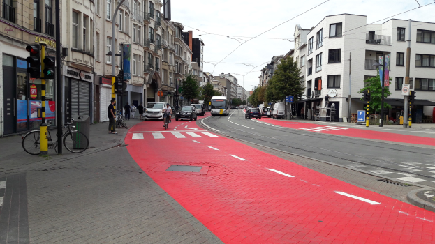 Rote Fahrradspur auf der Seite einer Stadtstraße mit Fahrzeugen, Fußgängern, Gebäuden, Bäumen und einem klaren blauen Himmel.