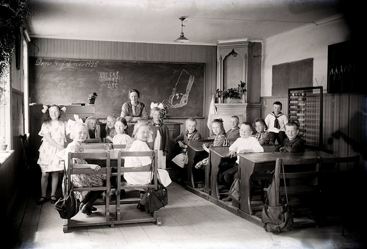 Schwarz-weißes Foto einer Klasse mit Kindern auf Bänken und einigen Stehenden, mit Taschen, Wandtafel mit Text, Topfpflanzen, Schrank, Fenster und Deckenbeleuchtung.