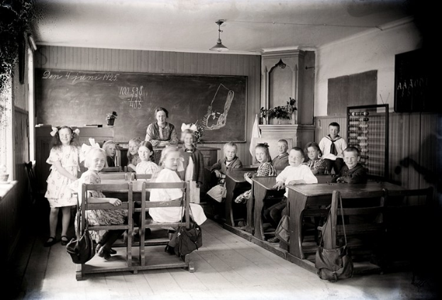 Schwarz-weißes Foto einer Klasse mit Kindern auf Bänken und einigen Stehenden, mit Taschen, Wandtafel mit Text, Topfpflanzen, Schrank, Fenster und Deckenbeleuchtung.