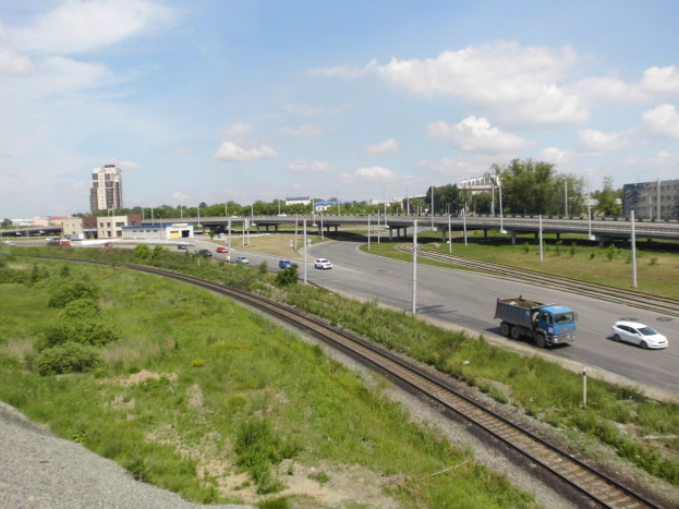 Eine Eisenbahnschiene verläuft durch eine Stadtlandschaft mit Fahrzeugen, Grünflächen, Polen, Brücken, Gebäuden und einem bewölkten Himmel.