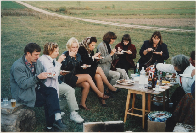Eine Gruppe von Menschen, die an einem Picknicktisch essen, mit Tellern, Flaschen, Gläsern und anderen Gegenständen auf einem grünen Rasen und einem blauen Himmel.