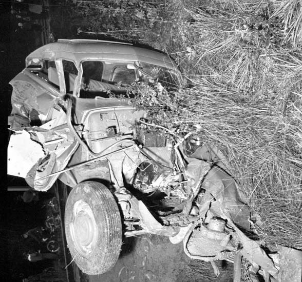 Schwarzes und weißes Foto eines stark beschädigten Autos mit eingedellter Front und zersplitterten Fenstern am Straßenrand, mit einem Baum und verstreuten Gegenständen im Hintergrund.