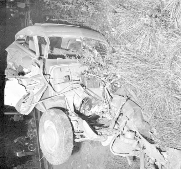Schwarzes und weißes Foto eines stark beschädigten Autos mit eingedellter Front und zersplitterten Fenstern am Straßenrand, mit einem Baum und verstreuten Gegenständen im Hintergrund.