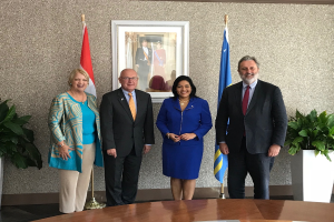 Eine Gruppe strahlender Menschen um einen Tisch mit Papieren versammelt, umgeben von Fahnen, Topfpflanzen und einem Foto auf dem Wand, feiert die Unterzeichnung eines Memorandums von Verständnis zwischen den Vereinigten Staaten und Kanada.