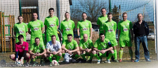 Eine Gruppe von Männern in grünen T-Shirts steht und hockt auf einem Fußballfeld mit einem Ball vor ihnen, ein Torpfosten rechts und ein Hintergrund von Bäumen, Gebäuden, Mästen, Fahrzeugen und dem Himmel.