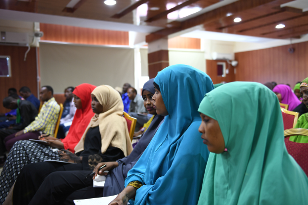 Eine Gruppe von Frauen sitzt in Stühlen in einem Raum, hält Bücher und Stifte in der Hand, mit einer Wand, Deckenleuchten und einer Klimaanlage im Hintergrund, an einer Workshop-Veranstaltung über Frauenrechte in Nigeria teil.