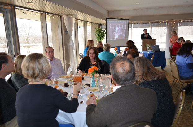 Eine Gruppe von Menschen, die um einen Tisch voller verschiedener Speisen sitzen, eine Person, die spricht, ein Projektor, der ein Bild anzeigt, eine Pflanze neben dem Projektor und ein Blick aus dem Fenster mit Vorhängen, Himmel und Bäumen im Hintergrund.
