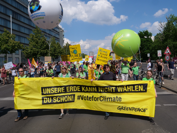 Eine Gruppe von Menschen marschiert mit einem gelben "W√§hle Klimaschutz"-Plakat, Ballons, Schildern, Fahrr√§dern, B√§umen, Laternenm√§sten und Geb√§uden im Hintergrund unter einem klaren blauen Himmel.
