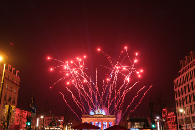 Eine Straßen Szenerie in Berlin an Silvester, gefüllt mit Gebäuden, Bäumen, Laternenpfählen, Verkehrszeichen, Zeltplanen und Menschen, mit einem Feuerwerk, das den Himmel erhellt.