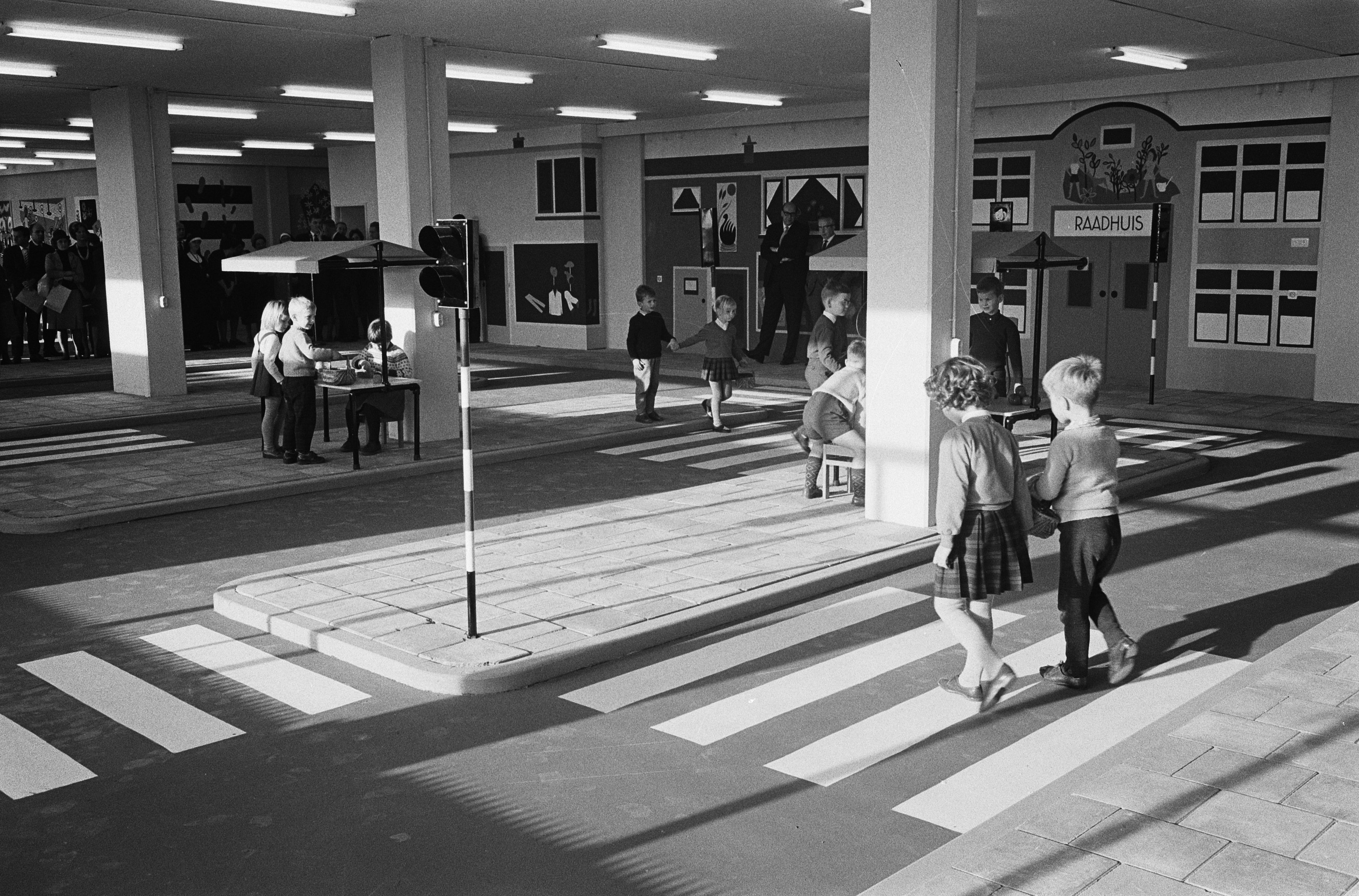 Schwarzes und weißes Foto von Kindern, die eine Straße an einer Ampel überqueren, mit Fußgängern in der Nähe, einem Verkehrssignal im Vordergrund und strukturellen Säulen mit Deckenleuchten im Hintergrund.
